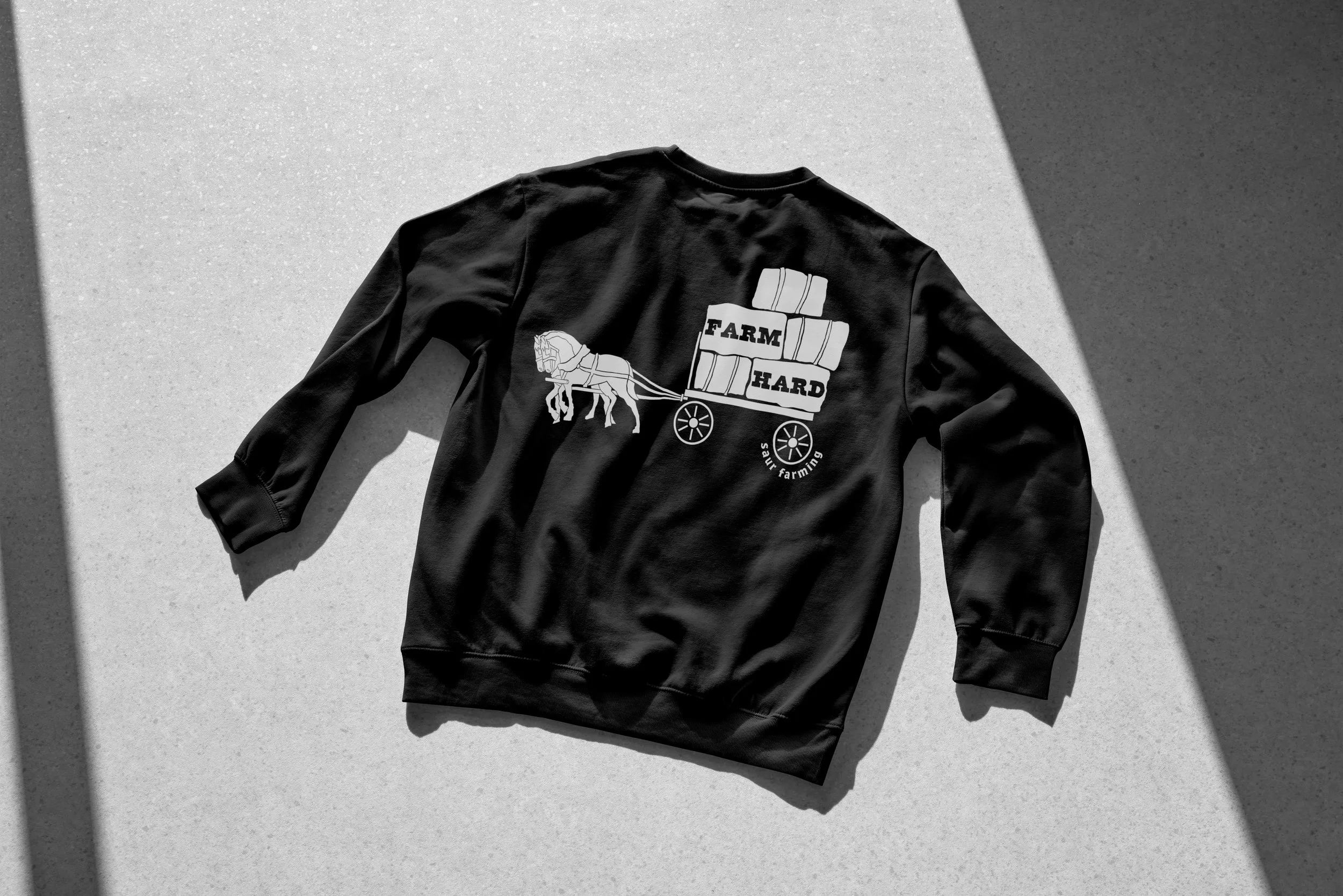 Black sweatshirt with a graphic of a horse pulling a cart loaded with hay bales and the words "FARM HARD" on the back, lying on a light-colored surface with shadows.