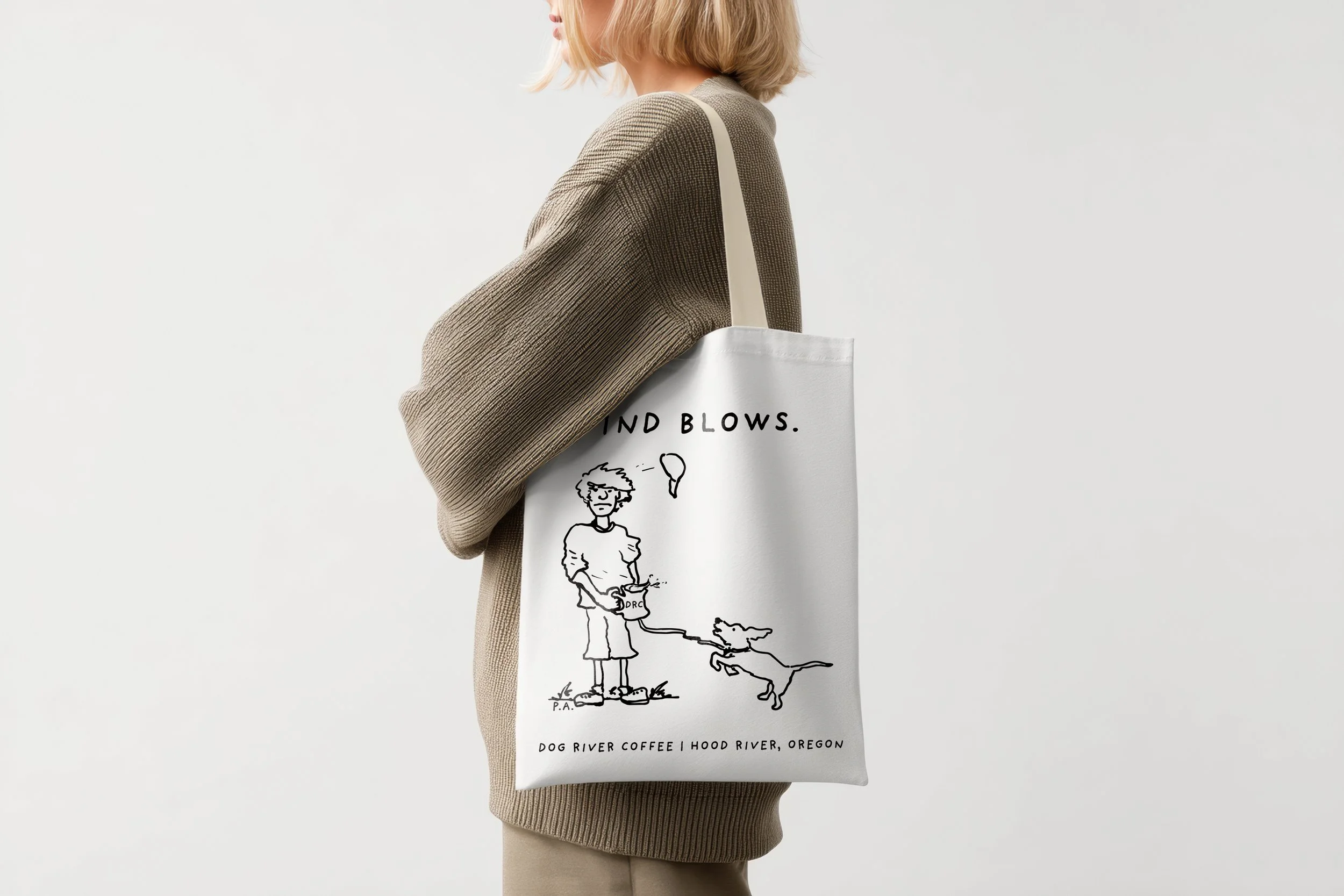 A woman wearing a beige sweater and pants holding a white tote bag with a black line drawing of a person walking a dog, and text reading "Dog Blows," "Dog River Coffee Hood River, Oregon."