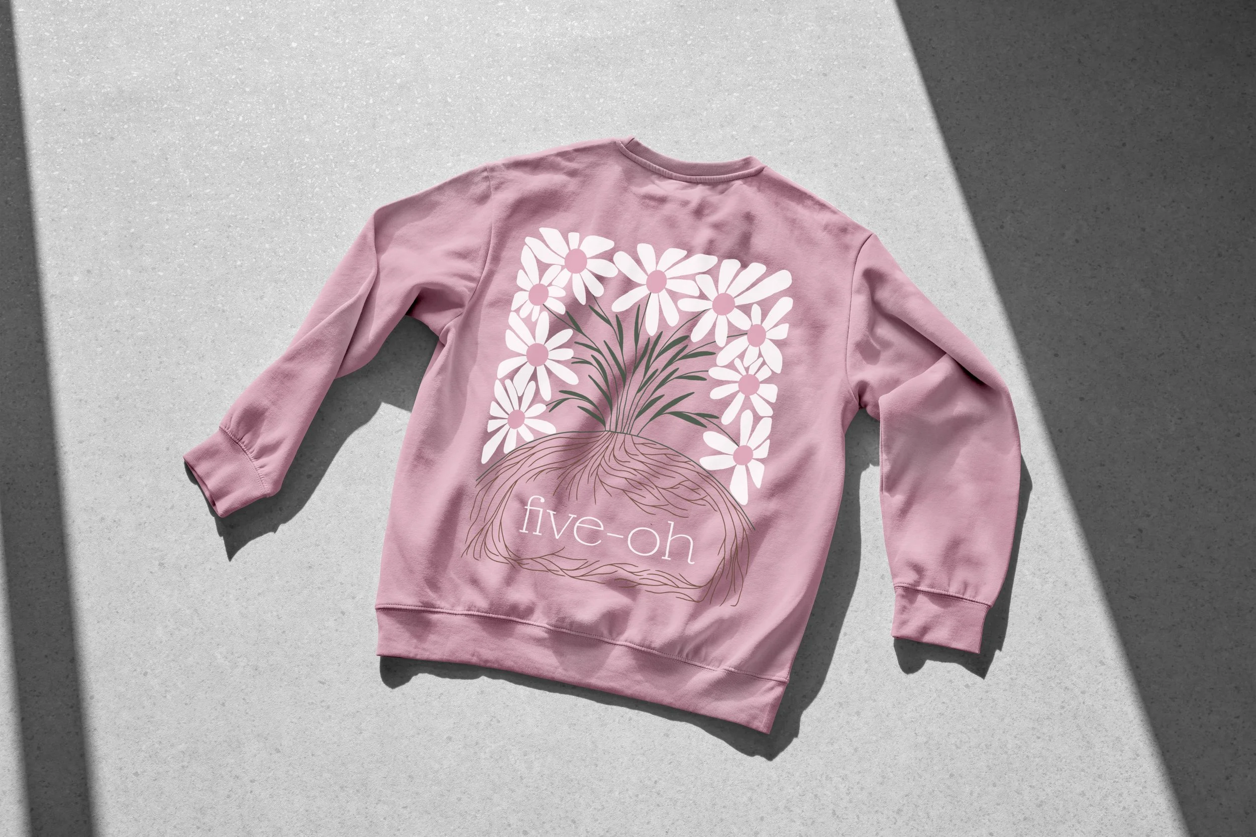 Pink sweatshirt with a graphic of white flowers and the text 'five-oh' on the back, lying on a light surface with shadows.
