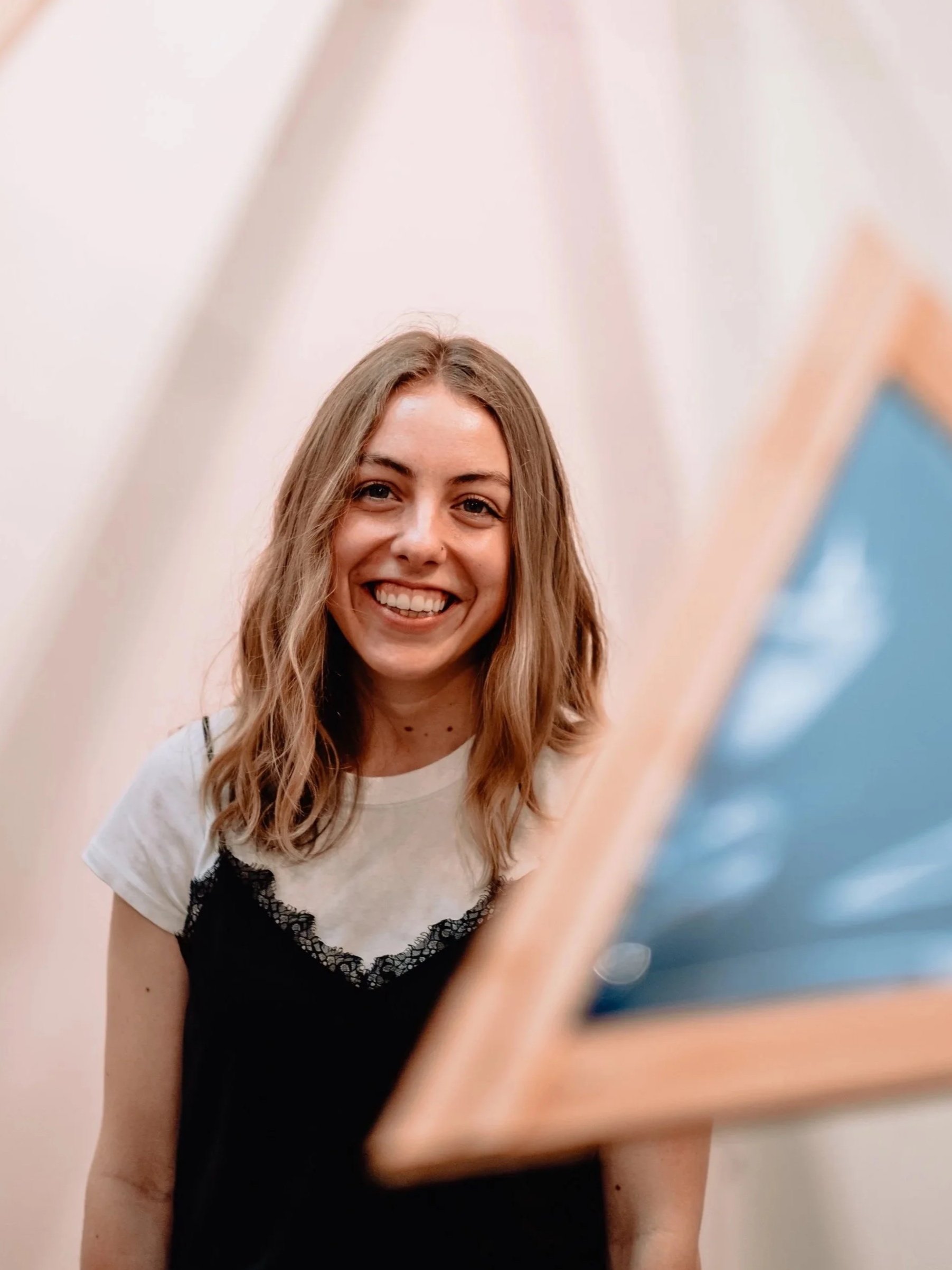 Young woman with shoulder-length wavy hair smiling, standing in front of a plain light-colored wall, with a blurred piece of art or photo frame partially visible in the foreground.