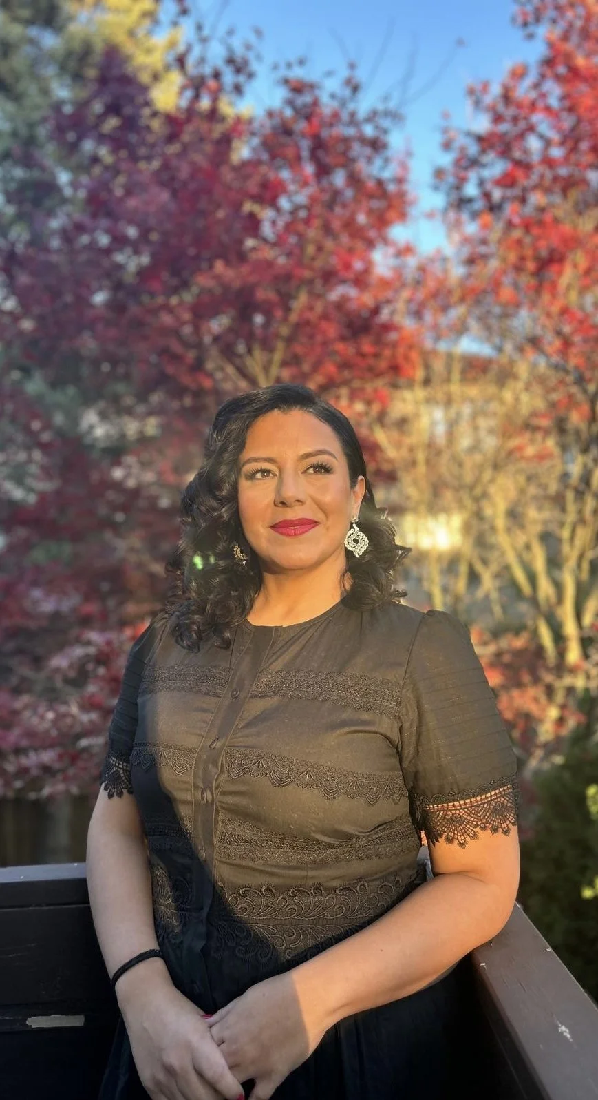 A woman with black wavy hair and red lipstick standing outdoors with autumn trees in the background.