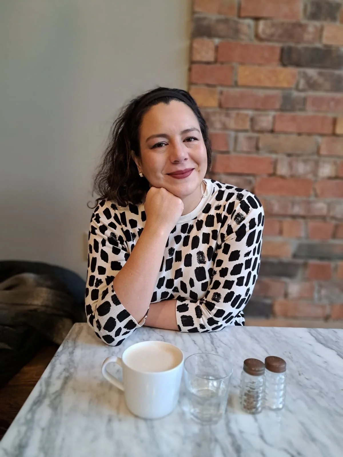 A woman with dark curly hair, wearing a black headband and a black and white animal print top, is sitting at a marble table with a white mug, a small glass of water, and two small salt and pepper shakers in front of her. She is smiling and resting her chin on her hand, with a brick wall in the background.
