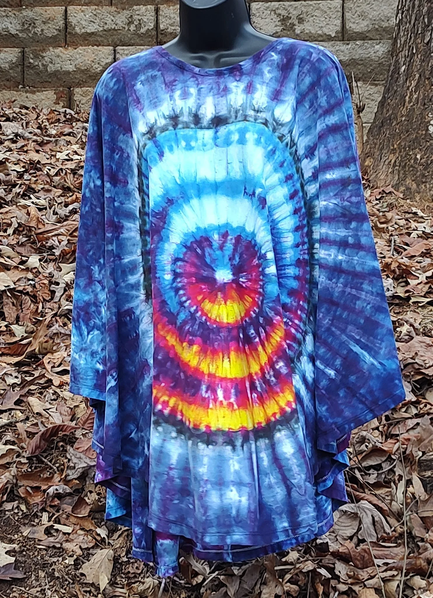A tie-dye circle poncho with a wigwag pattern in bright colors, including blue, purple, red, orange, and yellow, displayed outdoors in Fall.