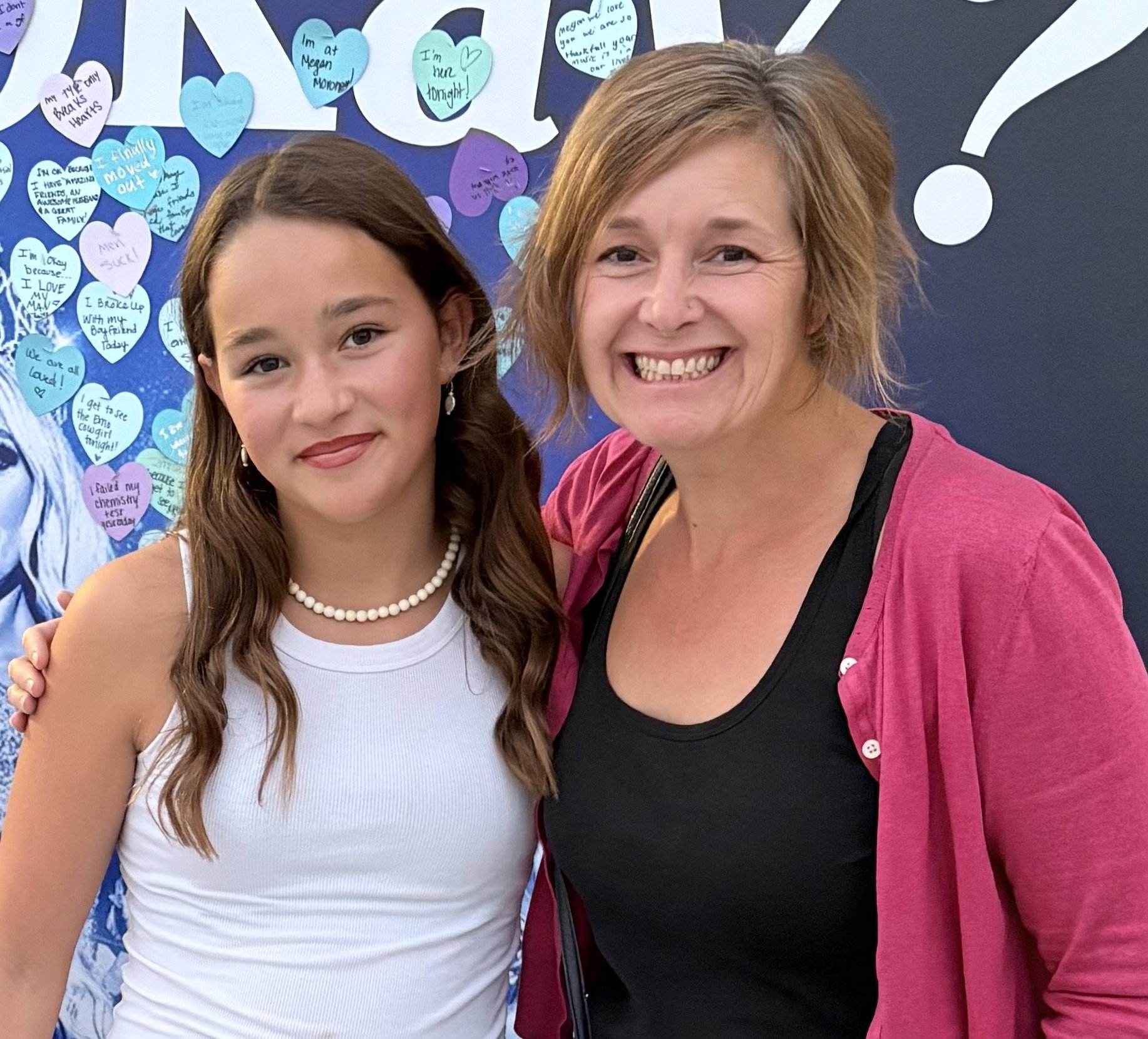 A girl and a woman smiling and posing together in front of a wall decorated with blue and purple heart-shaped notes and a large white question mark.