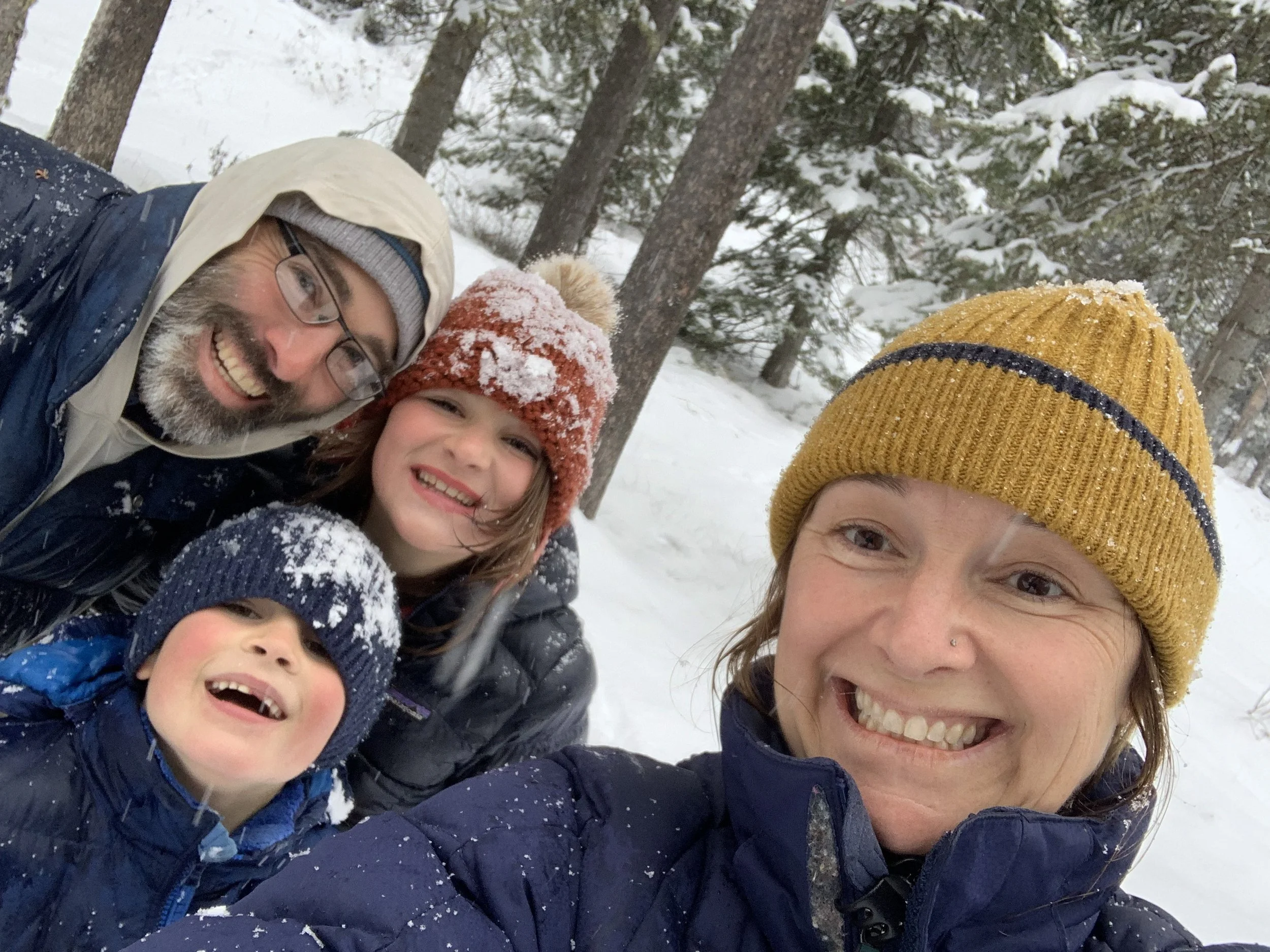 A group of five smiling people taking a selfie in a snowy forest. They are dressed in winter clothing, including hats and jackets, with snow on their hats and shoulders.