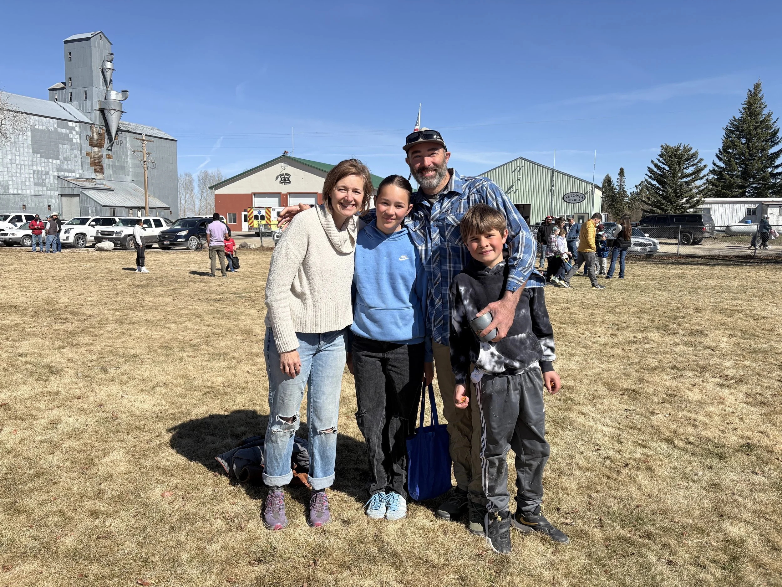A family of four, a woman, two children, and a man, standing outdoors on a grassy field with a clear blue sky, some buildings, and people in the background.