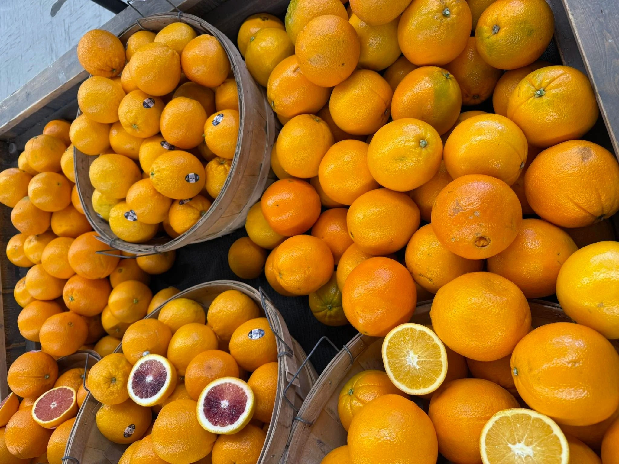 Pile of oranges and blood oranges at a market stall with some halved oranges showing inside. Some are in baskets and others are spread out on the surface.