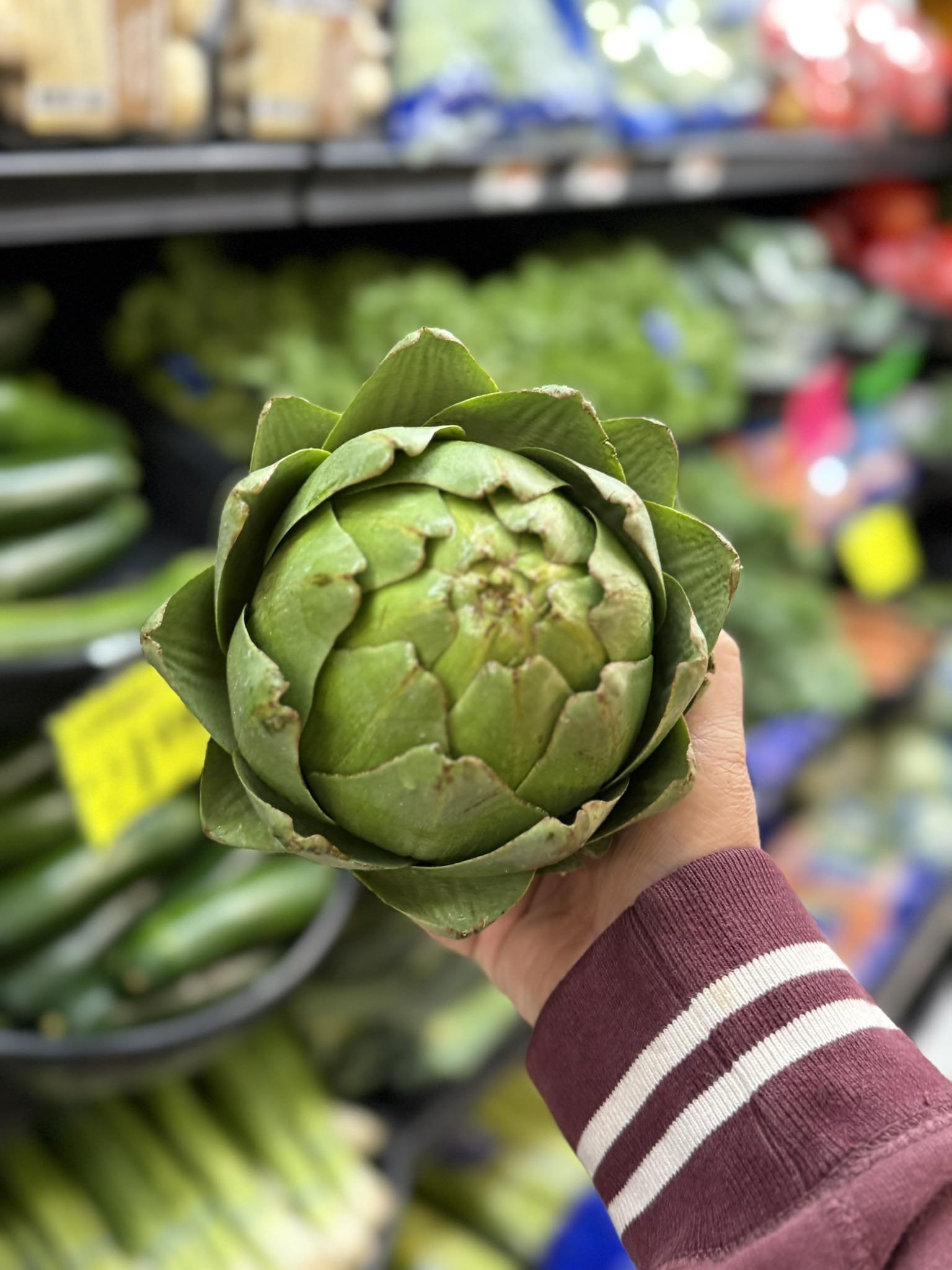 Person holding a fresh green artichoke in a grocery store aisle with other vegetables and produce in the background.