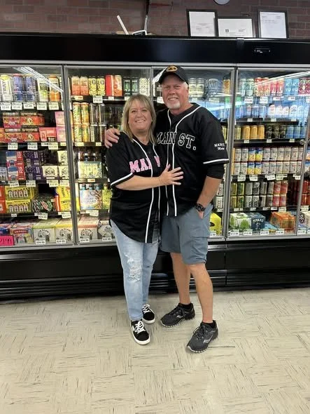 A smiling woman and man wearing matching black sports jerseys with white text, standing close together and hugging in front of a grocery store refrigerated section filled with dairy and other refrigerated products.