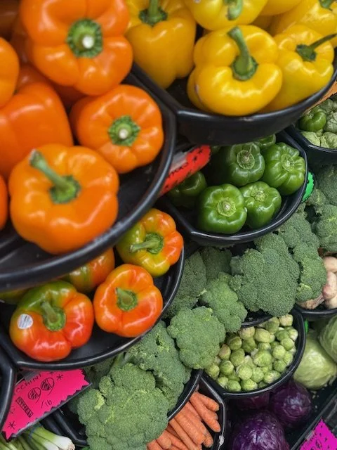 Assorted fresh bell peppers in orange, yellow, and green, broccoli, Brussels sprouts, carrots, and purple cabbage in a grocery store display.