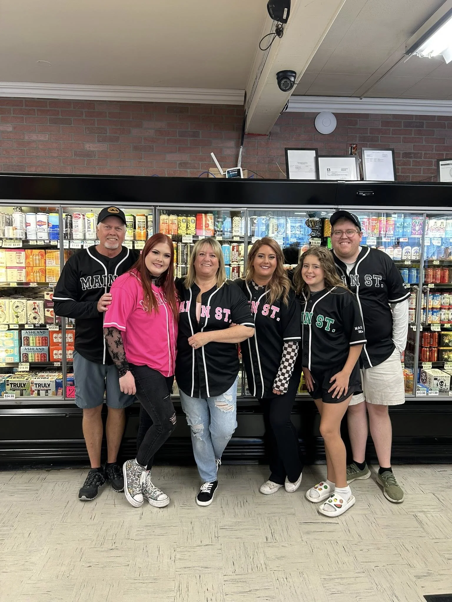 Group of six people standing in front of a refrigerated snack display, all wearing black baseball jerseys with white trim, some with the words 'Main St.' on them, inside a store with brick walls and tiled floors.