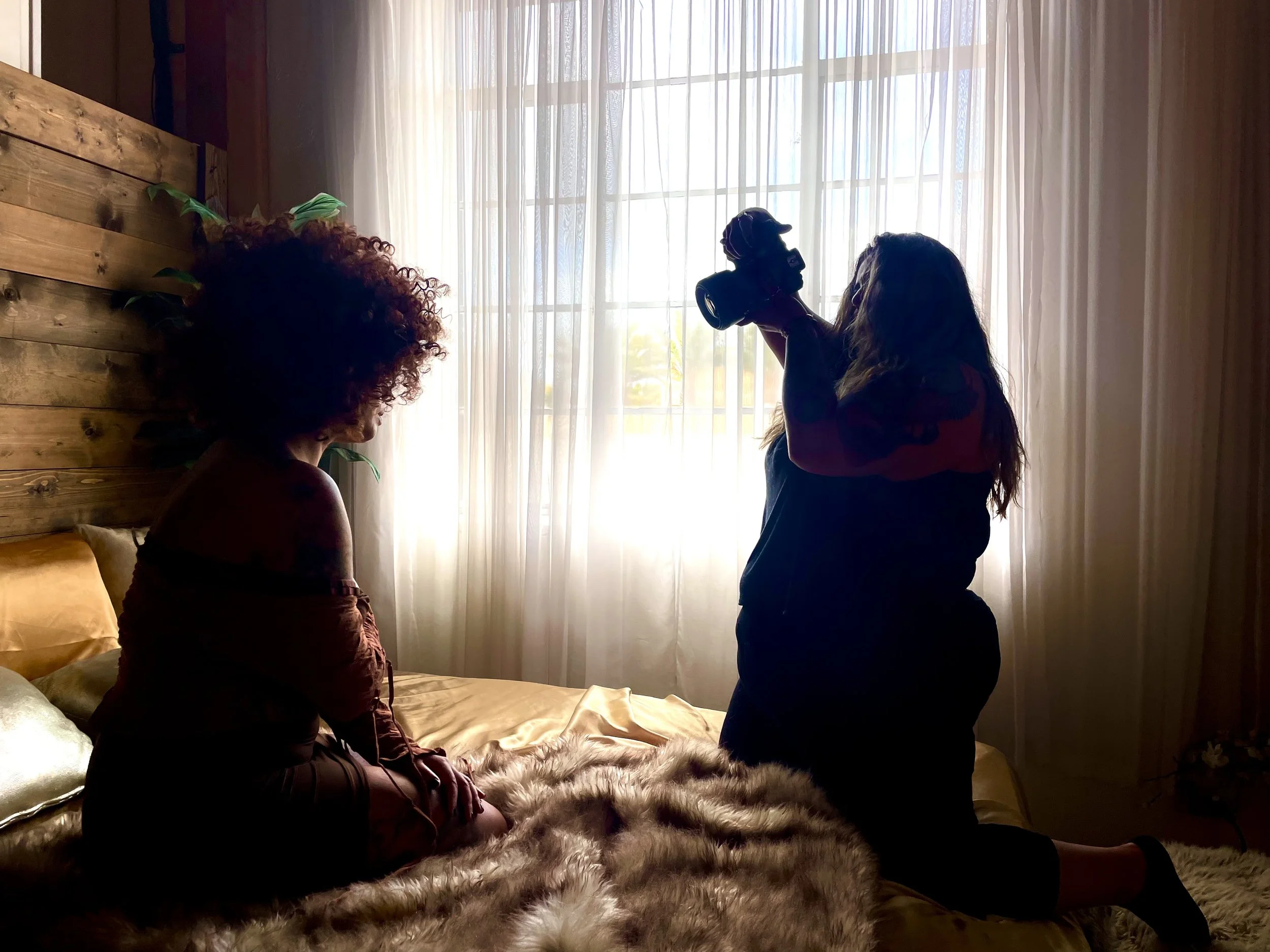 A woman with long hair holding a camera, a person with curly hair sitting on a bed with furry blanket, both in a room with sheer curtains and wooden wall.