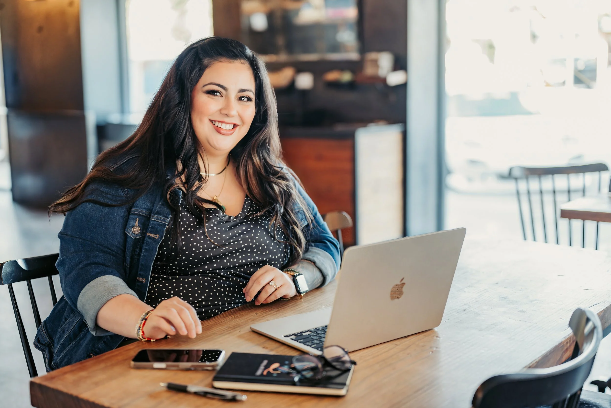 A woman with long dark hair smiling at a laptop in a cafe, sitting at a wooden table with a smartphone, glasses, a notebook, and a pen. Sunlight coming through the windows.
