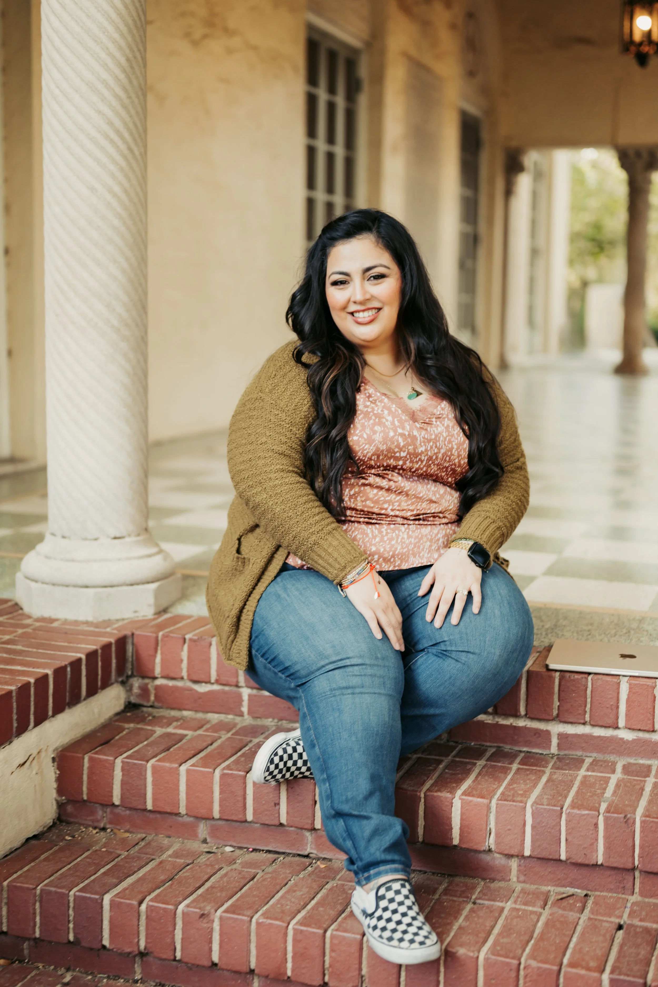 A woman sitting on brick steps outside a building, smiling, with long dark hair, wearing a brown cardigan, pink patterned shirt, blue jeans, checkered Vans sneakers, and accessories.