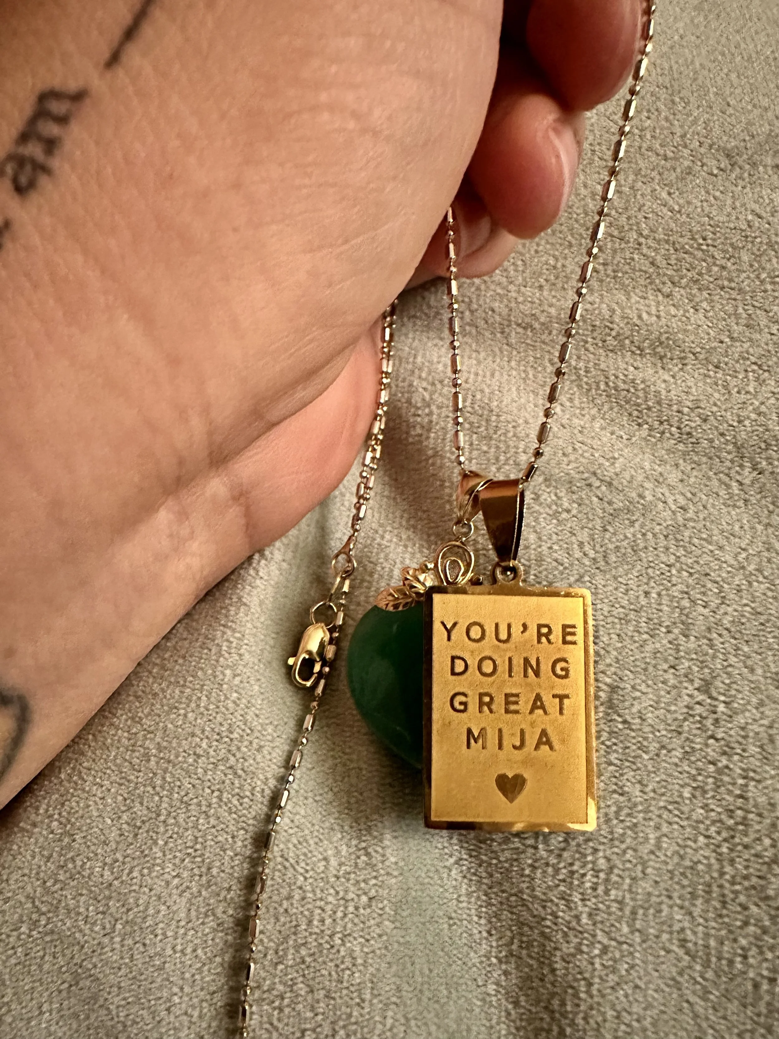 Close-up of a person holding a necklace with a pendant that says 'You're doing great Mija' and a gold heart at the bottom. The person's hand is partially visible with a tattoo on the wrist, and the background is a textured beige surface.