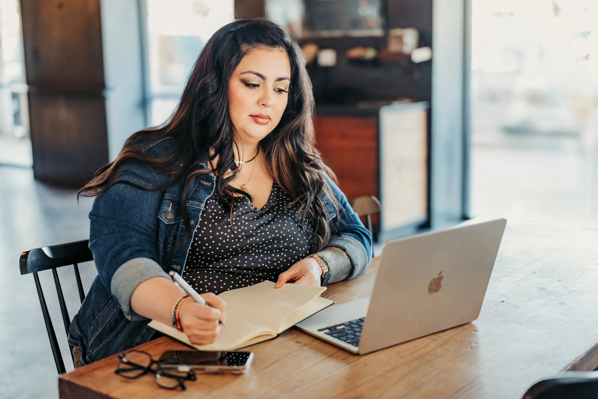 Young woman with long dark hair sitting at a wooden table, writing in a notebook with a laptop open in front of her, wearing a denim jacket and polka dot top, in a well-lit café or restaurant.