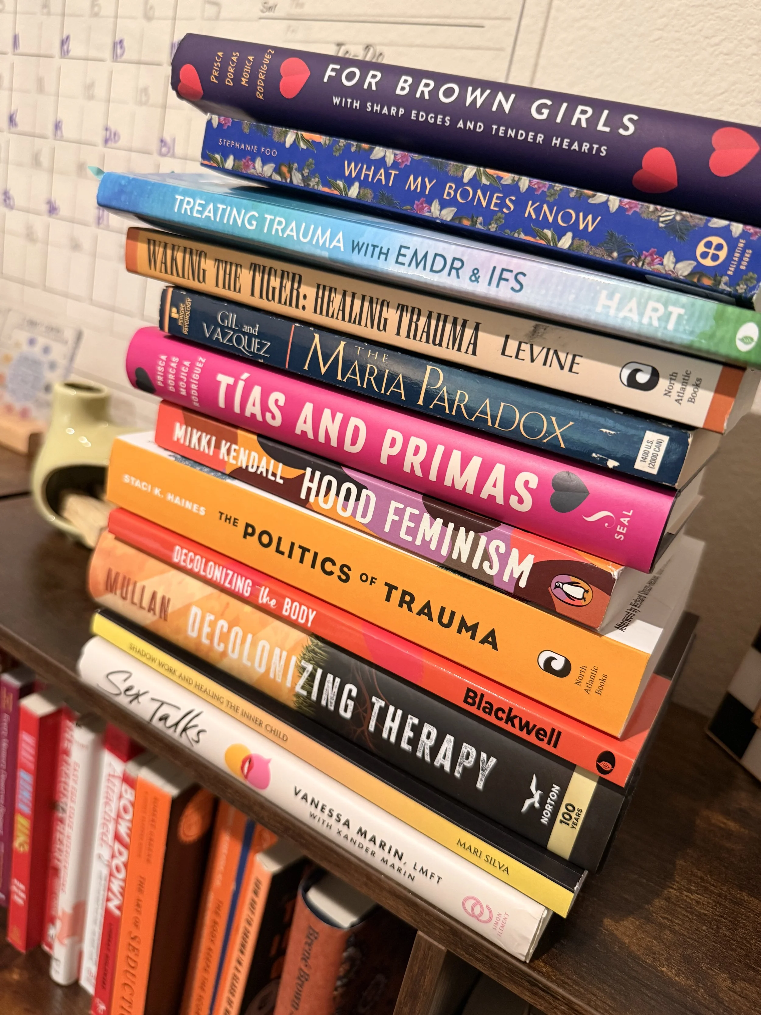 A stack of books on a bookshelf, with the titles including 'For Brawn Girls,' 'What My Bones Know,' 'Treating Trauma,' 'Waking the Tiger,' 'The Maria Paradox,' and others, with some books visible on the lower shelves.