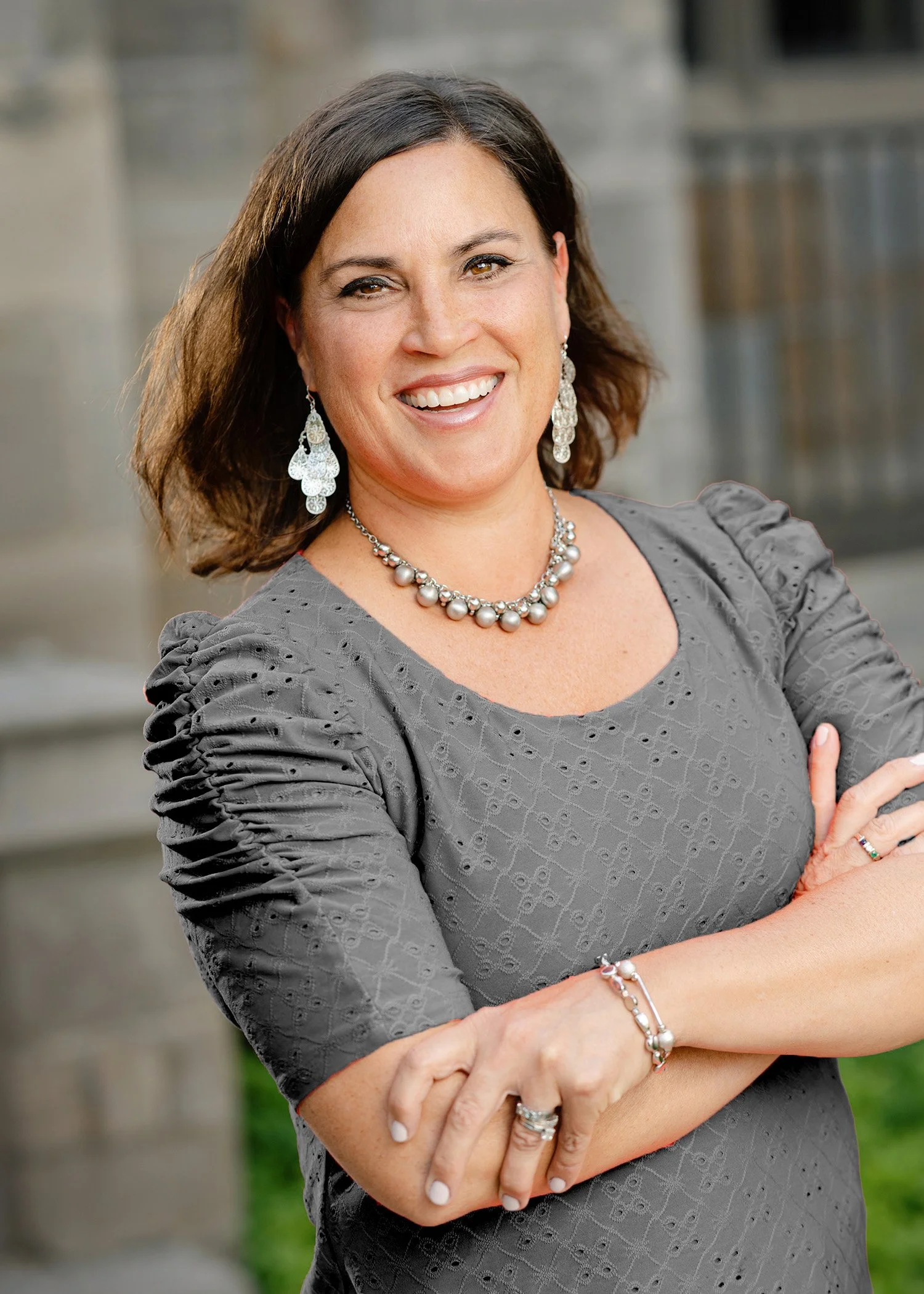 A smiling woman with dark brown hair in a grey, textured dress, wearing silver jewelry, standing outdoors with arms crossed.