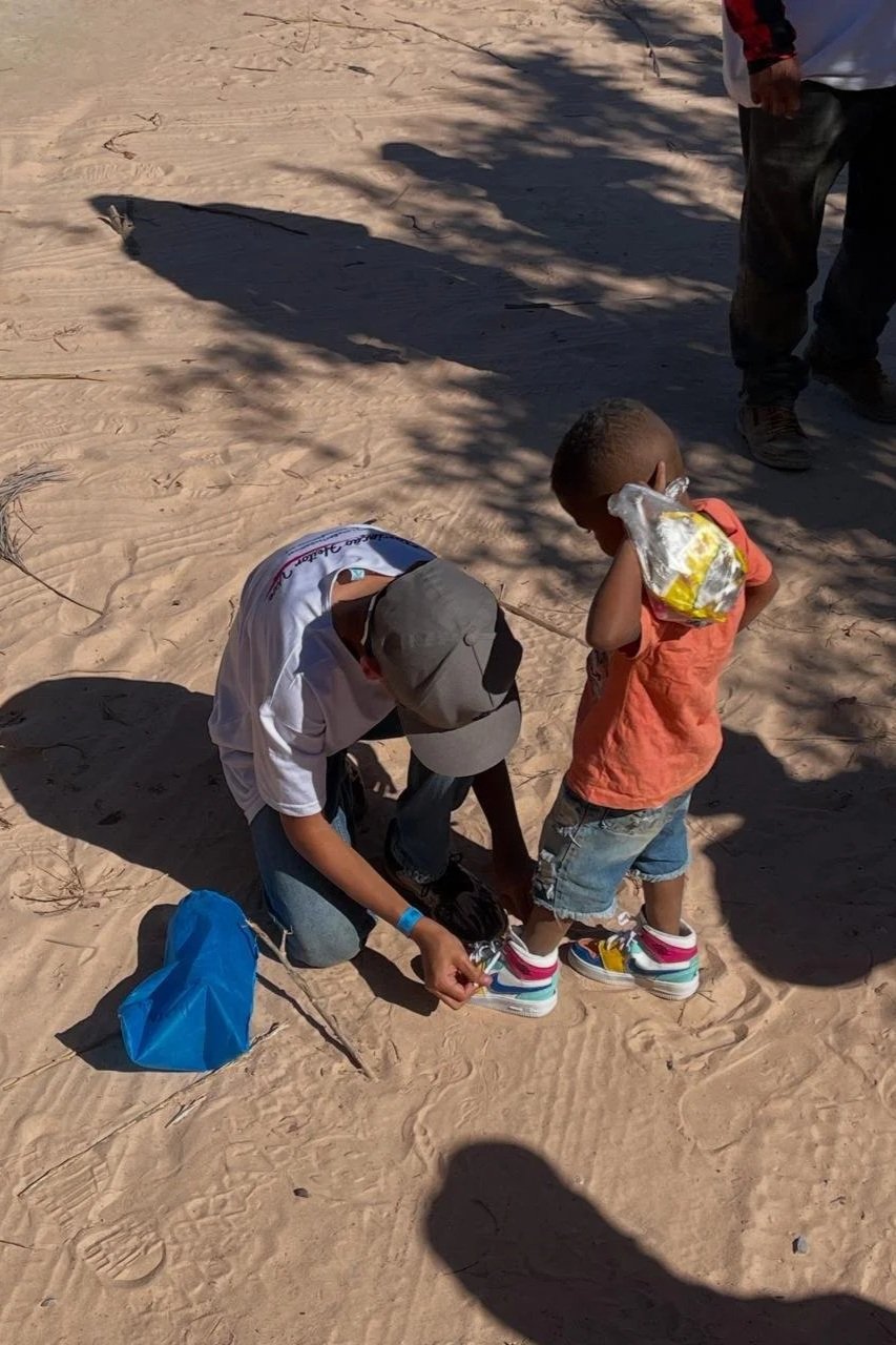 Criança recebendo ajuda de um adulto para calçar tênis em uma área de areia, com sombra de árvores ao redor.