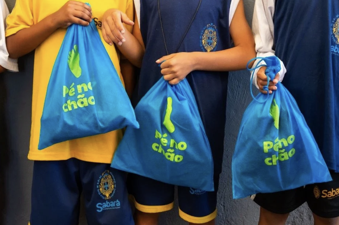 Crianças vestidas com uniformes escolares da escola Sabará, segurando sacolas azuis com a frase 'Pé no chão' em letras verdes.