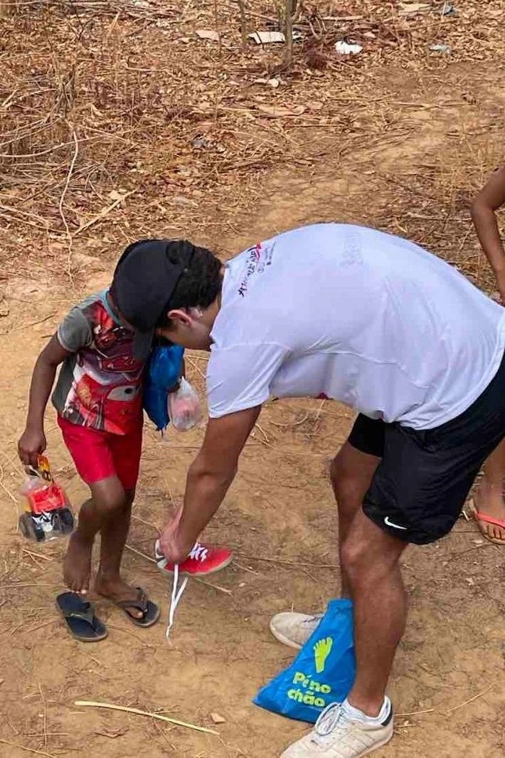 Homem ajudando uma criança a colocar o tênis, ambos na rua de terra, com roupas casuais e objetos nas mãos.