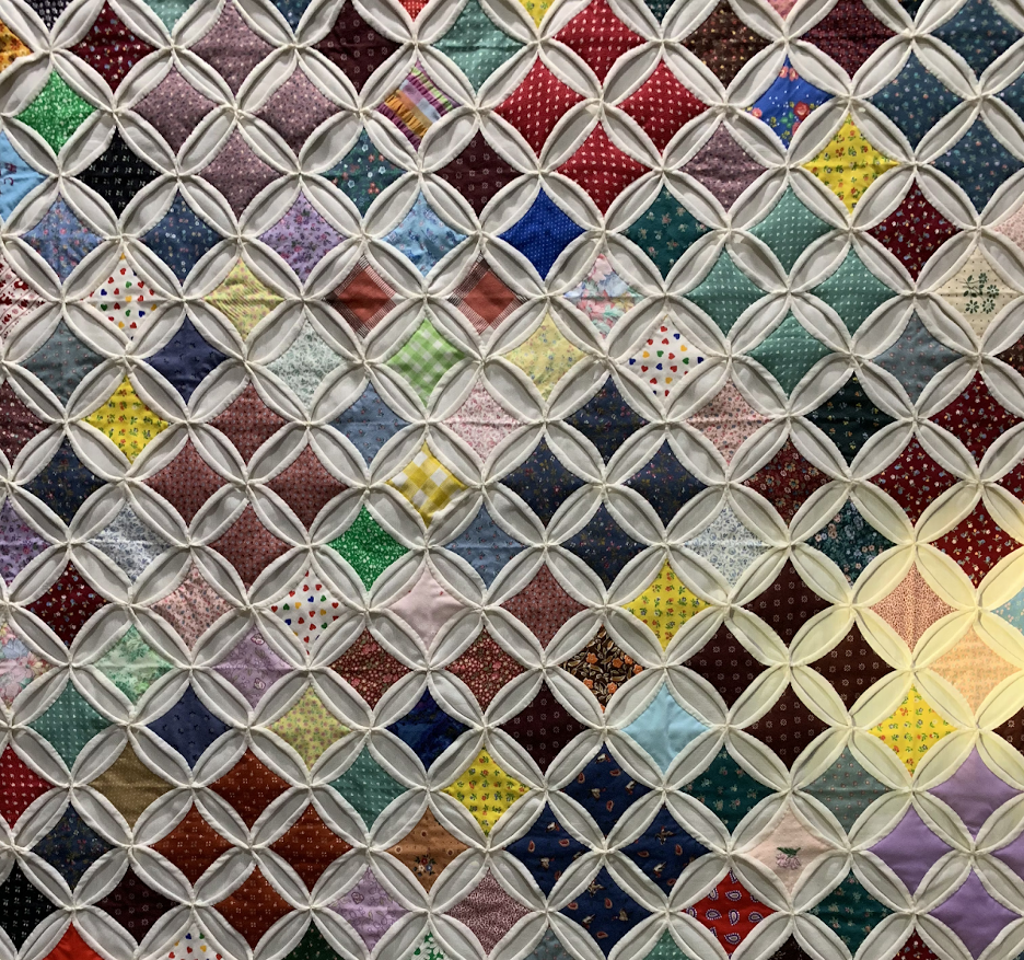 Detalhe de uma colcha de retalhos formada por pequenos quadrados de tecido colorido, protegidos por uma rede de elástico branco formando círculos entre os quadrados.