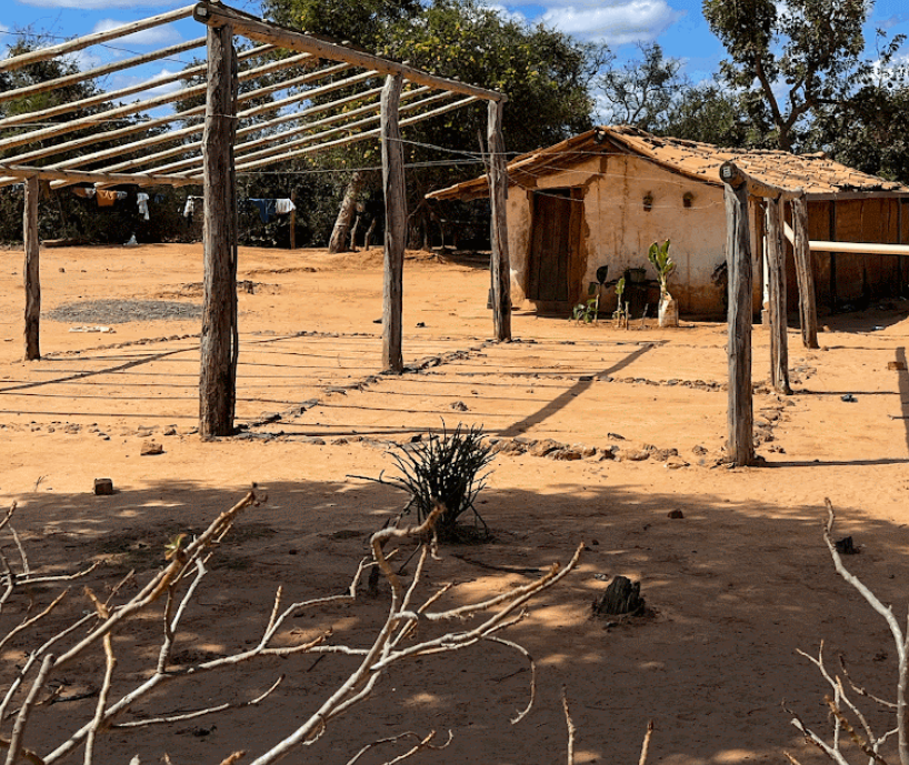 Casa de barro com telhado de madeira e área cercada com postes de madeira e cordas, em terreno arenoso com árvores ao fundo.