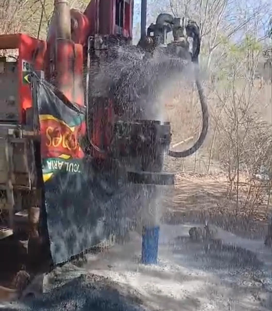 Caminhão despejando água em uma estrada de terra, com árvores secas ao fundo.