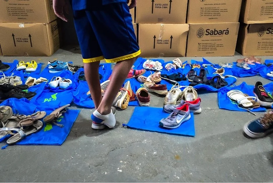 Vários sapatos e tênis espalhados sobre tapetes azuis, com uma pessoa de camisete azul e shorts ao fundo e caixas de papelão ao fundo.