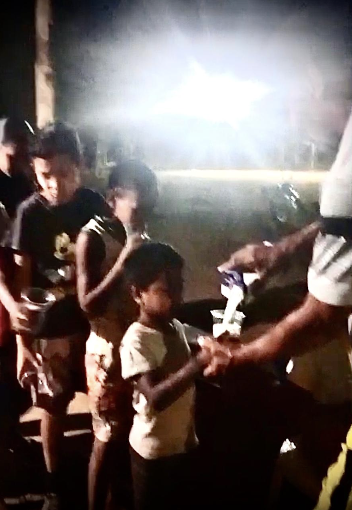 Crianças e adultos em uma rua à noite, com uma pessoa entregando algo a uma criança.