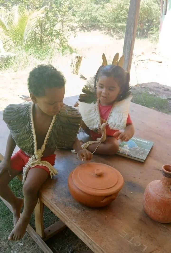 Crianças indígenas vestidas com roupas tradicionais, uma delas usando uma touca feita de penas, brincando com uma panela de barro sobre uma mesa de madeira ao ar livre.
