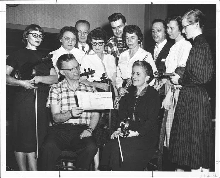 Group of nine women and two men indoors, with some holding violins and recorders, one woman seated with a violin, and a man seated holding a paper, possibly in a music or school setting.