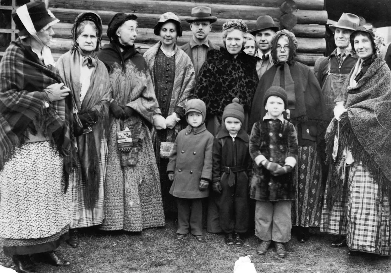 Black and white photo of a large family outside, dressed in old-fashioned clothing. Adults and children are standing together in front of a log cabin, some holding hands, with serious expressions.