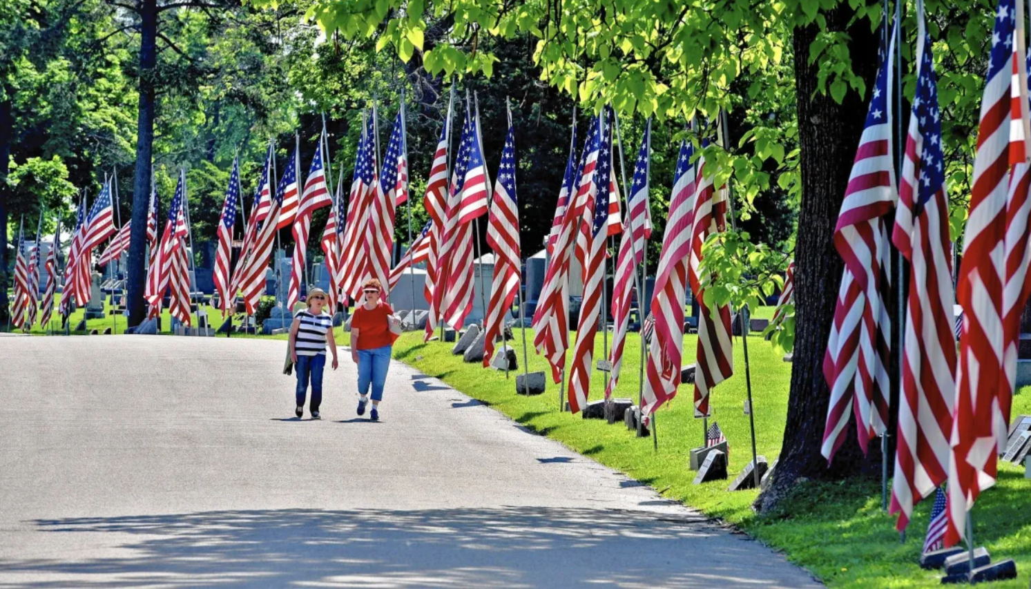 A memorial with numerous American flags and two women walking along a paved path, surrounded by tall trees and green grass.