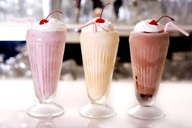 Three milkshake glasses with whipped cream and cherries on top, each with a straw, sitting on a white surface.