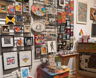 A wall display of various framed artworks, crafts, and decorations, with a small table and a rainbow flag on a wooden cabinet.