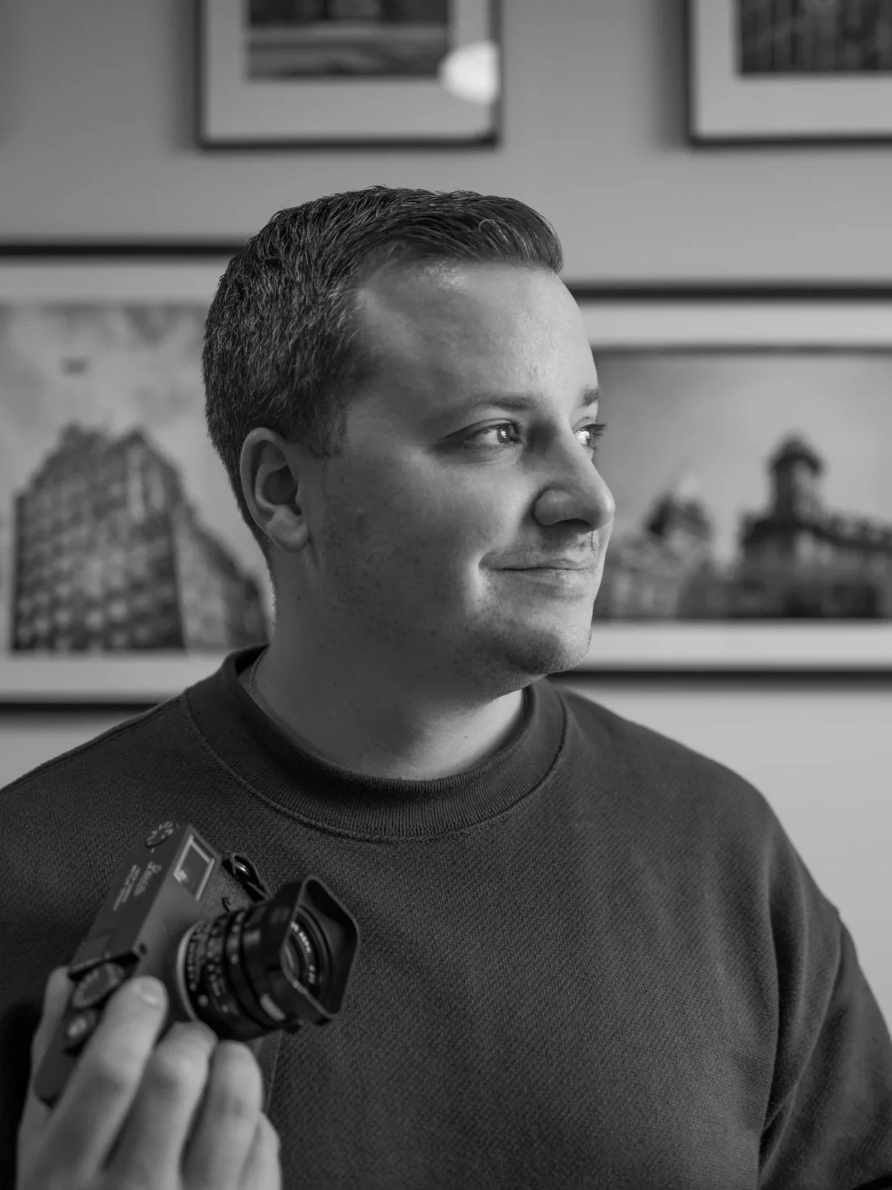 A young man with short, dark hair looking to the side, holding a vintage camera in his right hand, with framed cityscape photos hanging on the wall behind him.