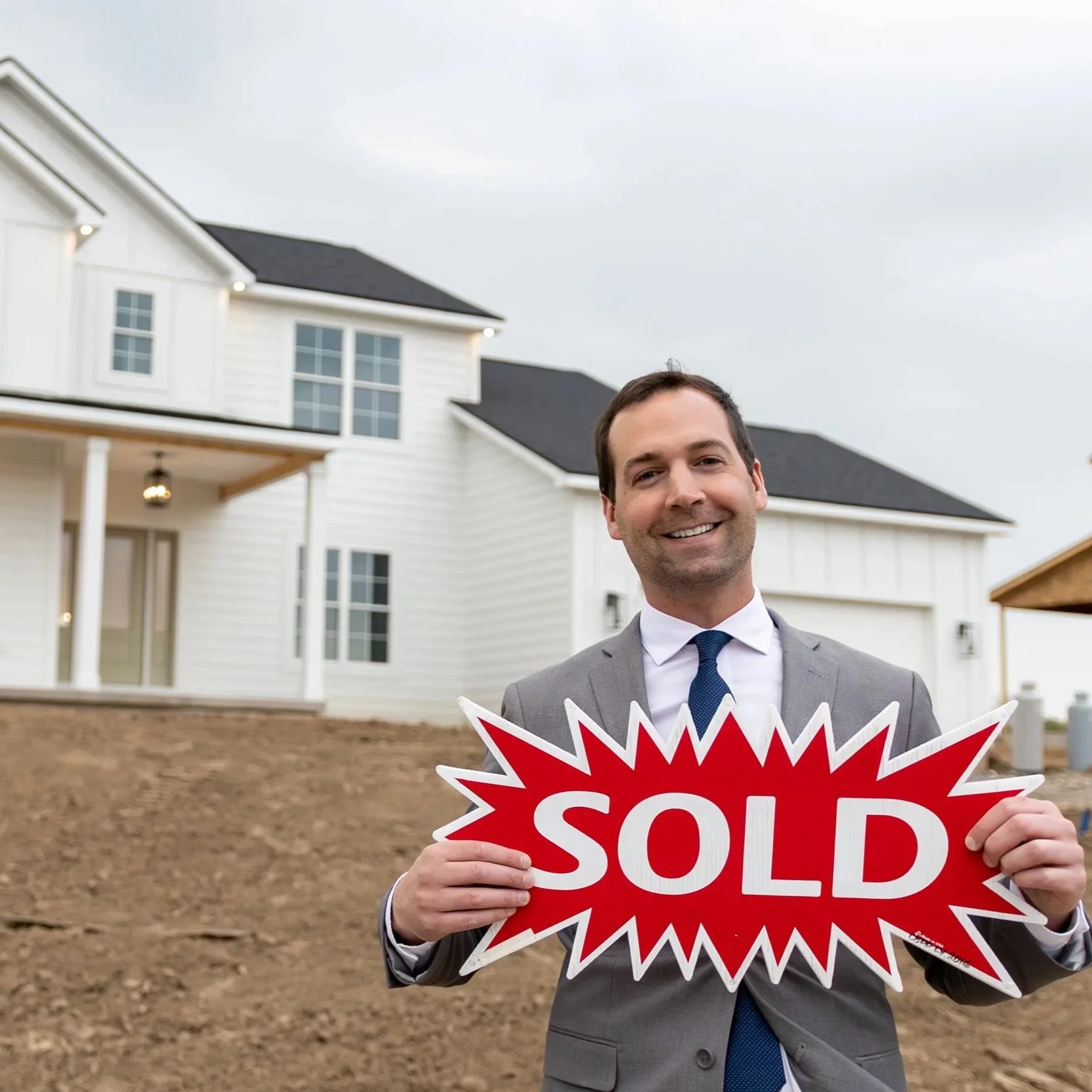 A man in a gray suit holding a red and white 'SOLD' sign in front of a house with a white exterior, multiple windows, and a dark roof.