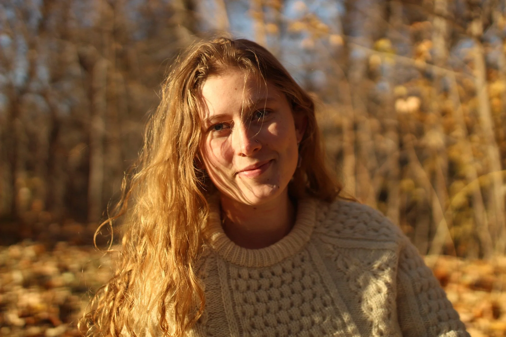 A young woman with long, wavy red hair smiling outdoors during autumn, wearing a beige knitted sweater, with trees and fallen leaves in the background.