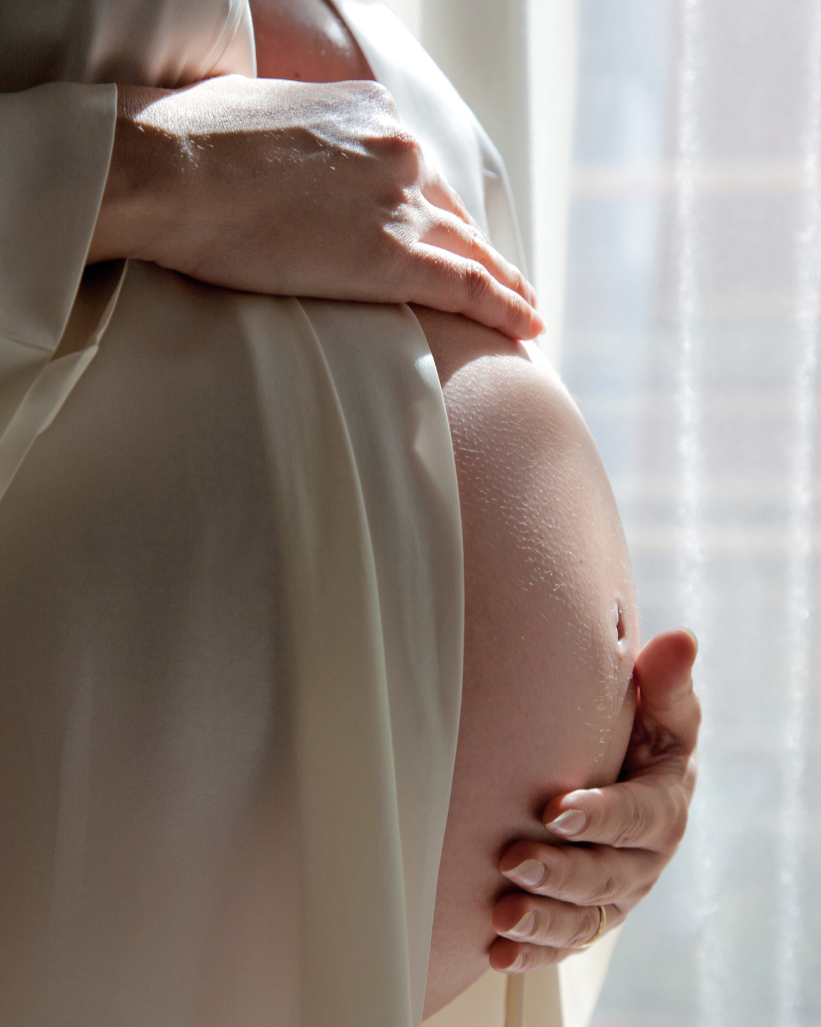 Close-up of a pregnant woman holding her bare belly with both hands, standing near a window with soft natural light.
