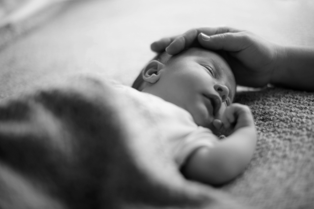 A baby sleeping on a textured surface with an adult hand gently resting on their head.