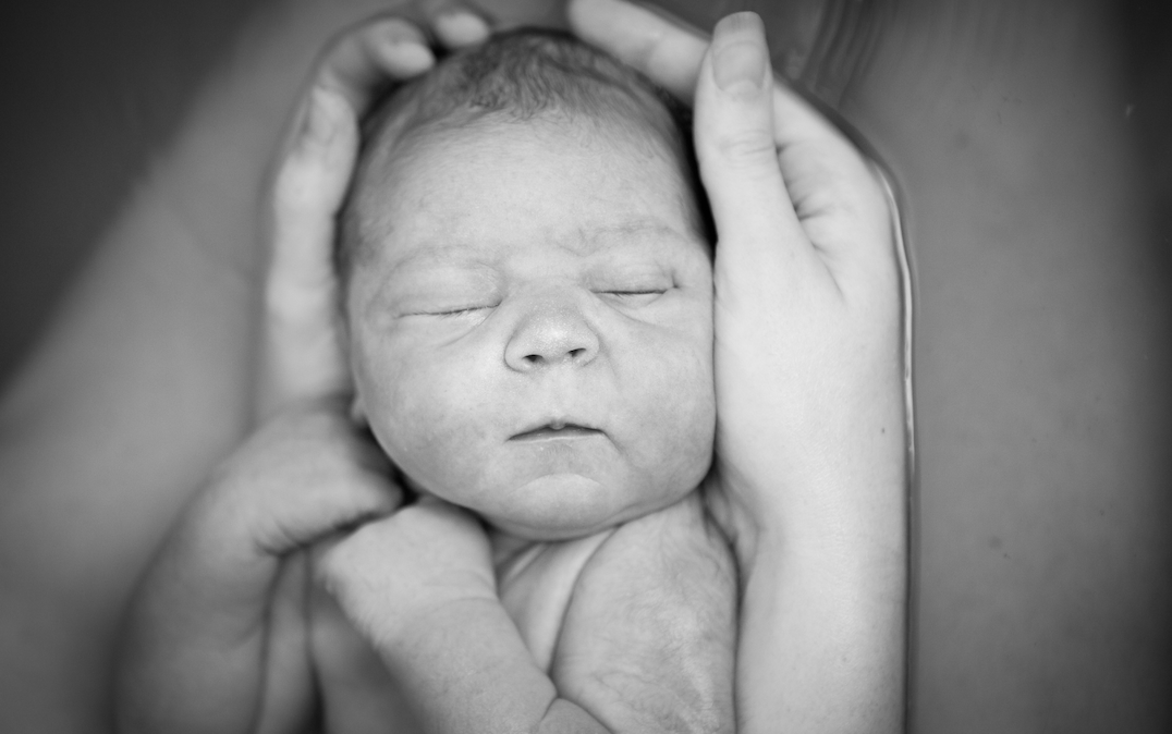 Black and white photo of a newborn baby sleeping peacefully, being gently held by adult hands.