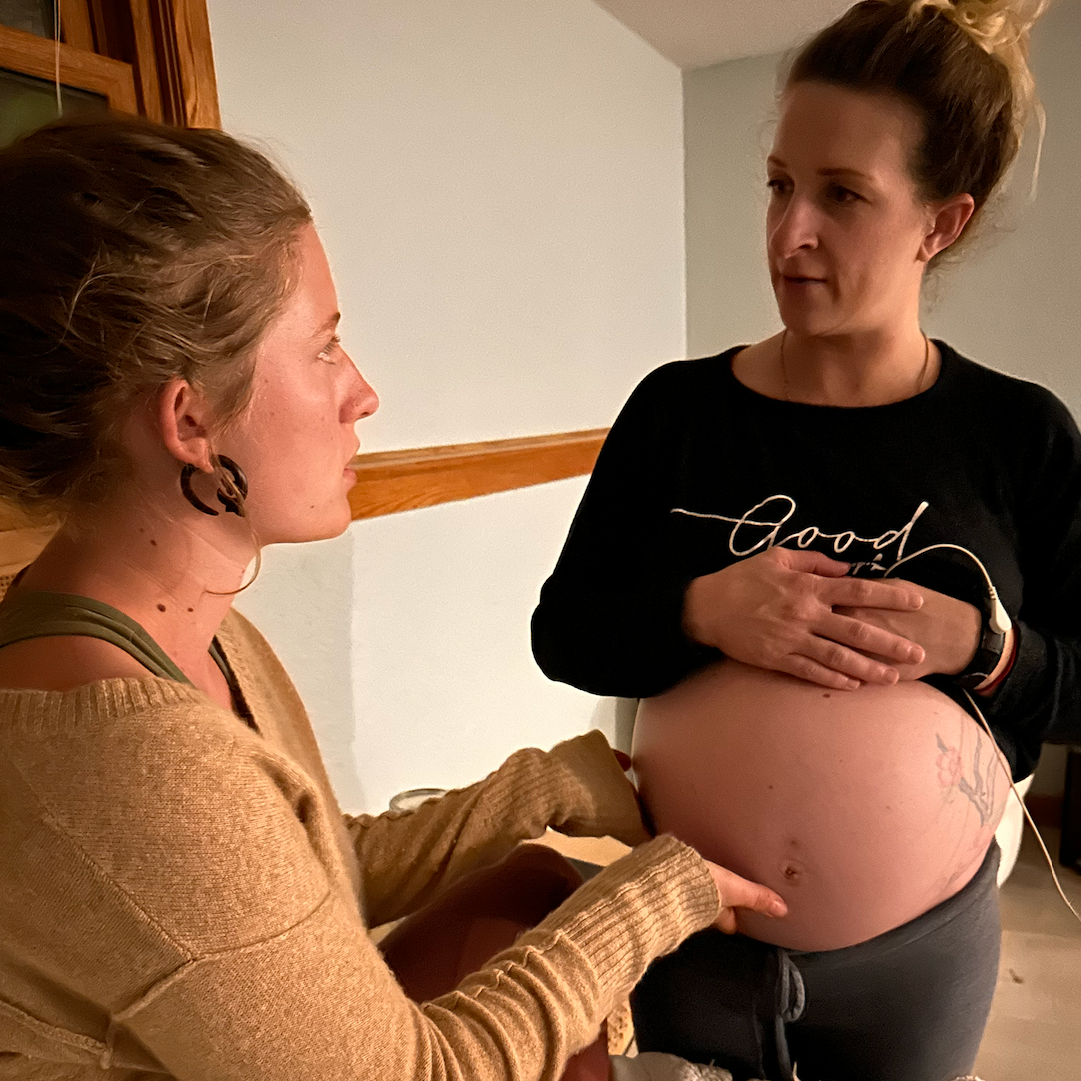 A pregnant woman standing and holding her belly while talking to another woman who is seated, pointing at her belly, in a home setting.