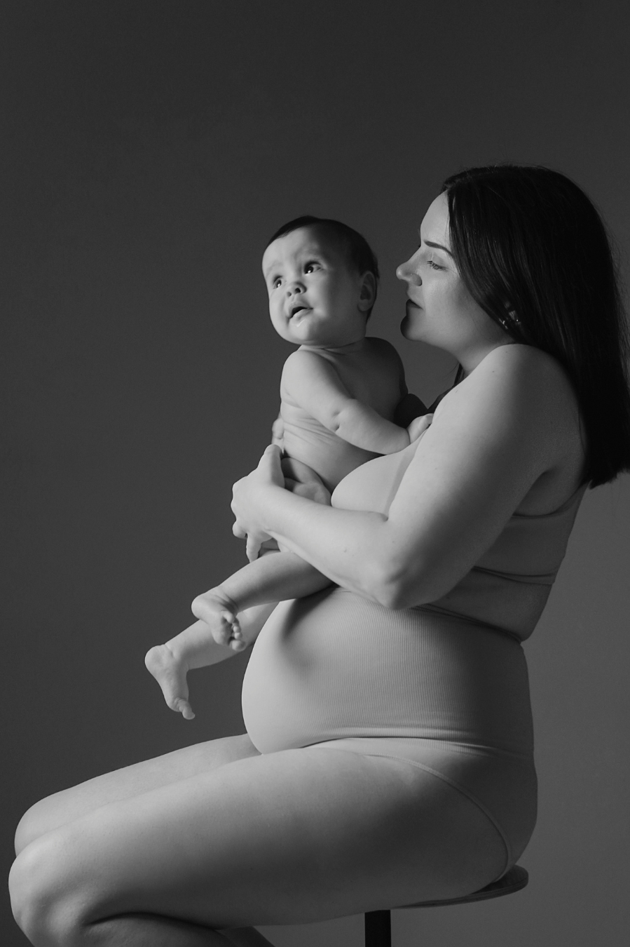 Black and white photo of a woman holding a baby, sitting on a stool against a dark background.