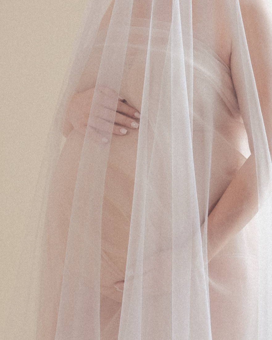 A nude person covered partially by sheer white fabric, with one hand covering their chest, standing against a light beige background.