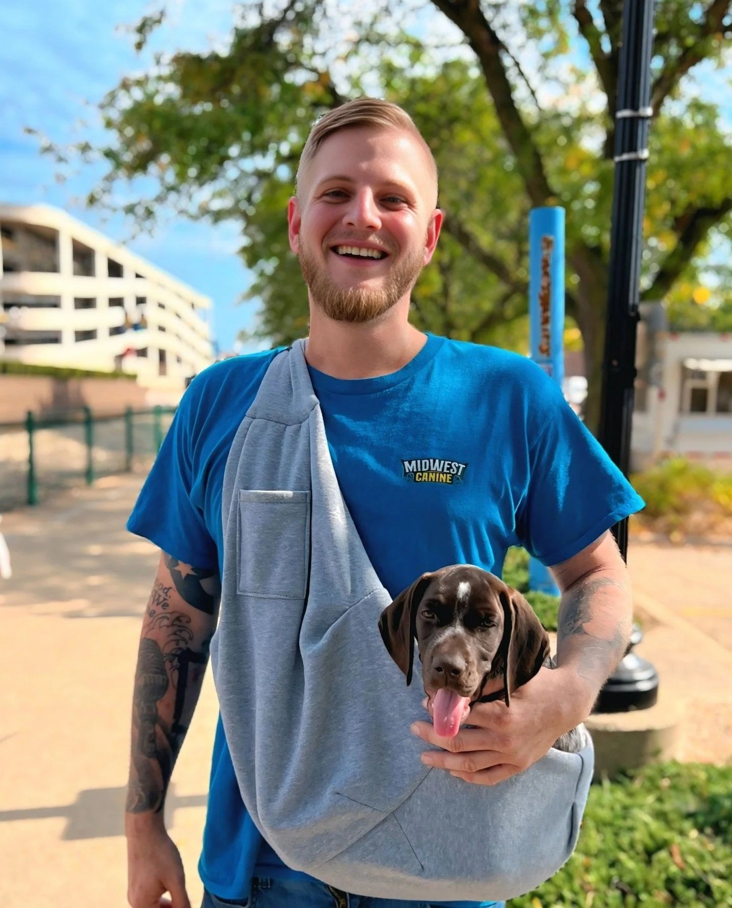 A smiling man wearing a blue t-shirt with 'Midwest Canine' on it, holding a brown puppy with its tongue out, outdoors on a sunny day with trees and buildings in the background.