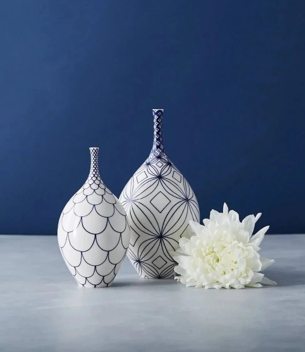 Two decorative white vases with black geometric patterns and a white flower on light-colored surface against a dark blue background.