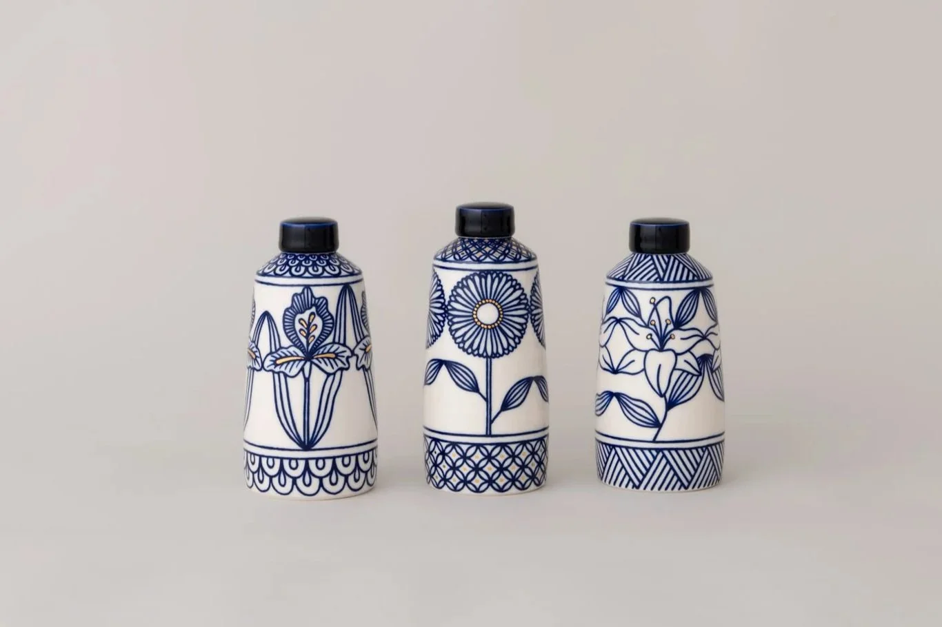 Three ceramic bottles with blue floral and geometric patterns, black caps, against a plain white background.