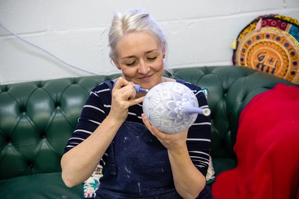 A woman with gray hair sits on a green tufted leather couch, painting intricate designs on a round ceramic object with a small brush. She wears a striped shirt and has a gentle smile. Behind her, colorful decorated items are visible, including a vibrant tapestry or cushion.