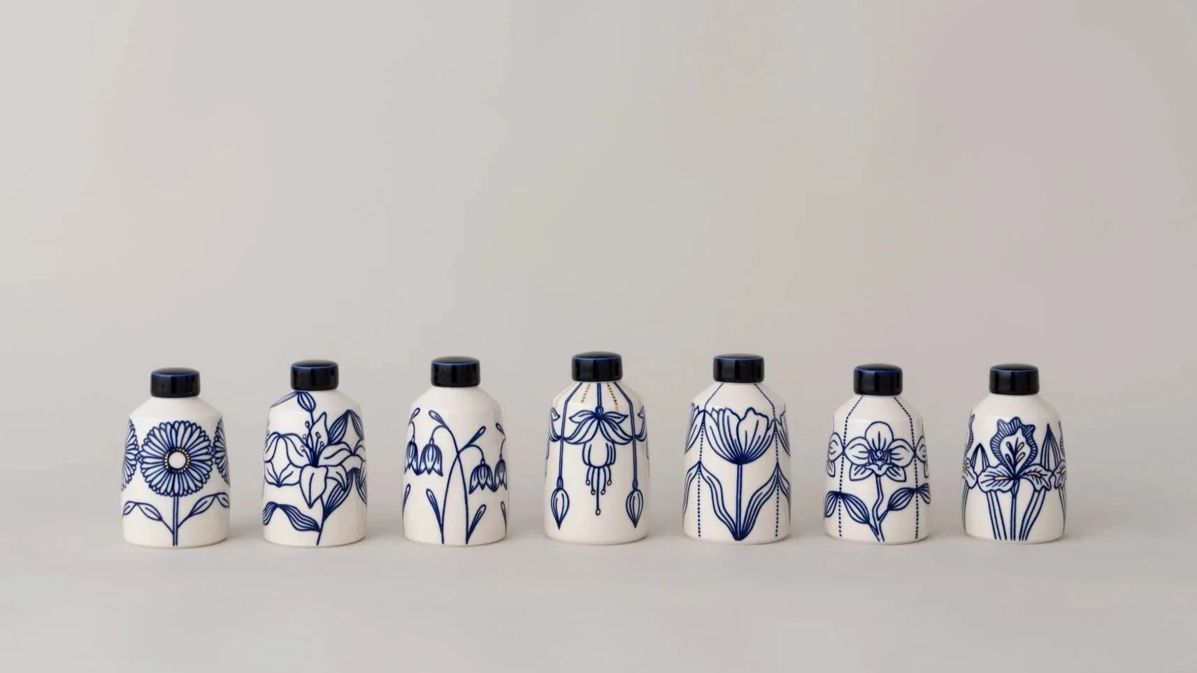 Seven white bottles with black caps, each decorated with blue floral patterns, arranged in a row on a white background.